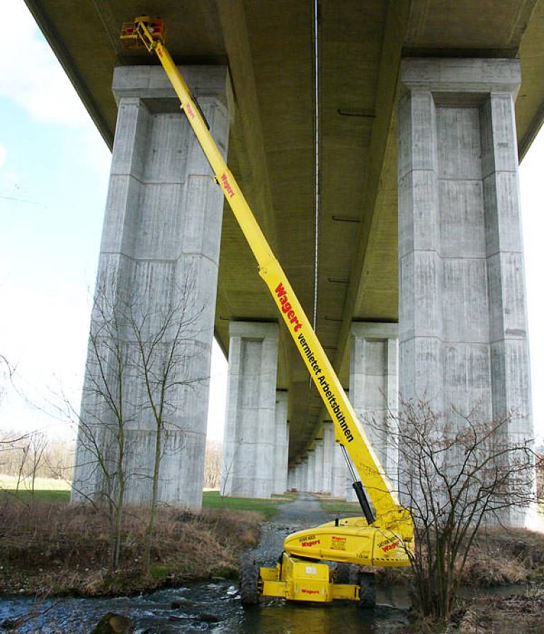 Prüfungsarbeiten an der Unterseite einer Autobahnbrücke, wobei die Wagert Teleskoparbeitsbühne in einem flachen Bach unter der Autobahnbrücke steht.